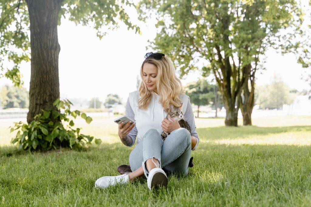 a woman sitting on the grass while using her phone and holding her cat