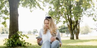 a woman sitting on the grass while using her phone and holding her cat